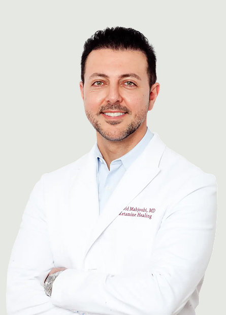 A man with short dark hair and a trimmed beard, wearing a white lab coat and light blue shirt, stands with arms crossed and smiles at the camera.