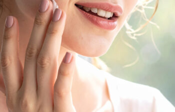 Close-up of a person with a slight smile applying a dab of white moisturizer to their cheek with their fingertips.